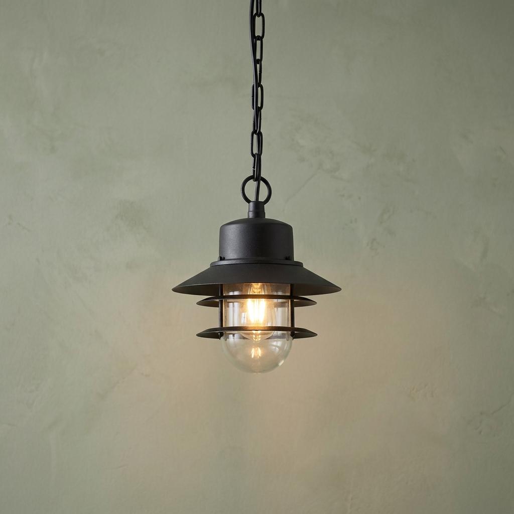 Black industrial-style outdoor chain lantern with clear glass shade illuminated against a green textured wall.