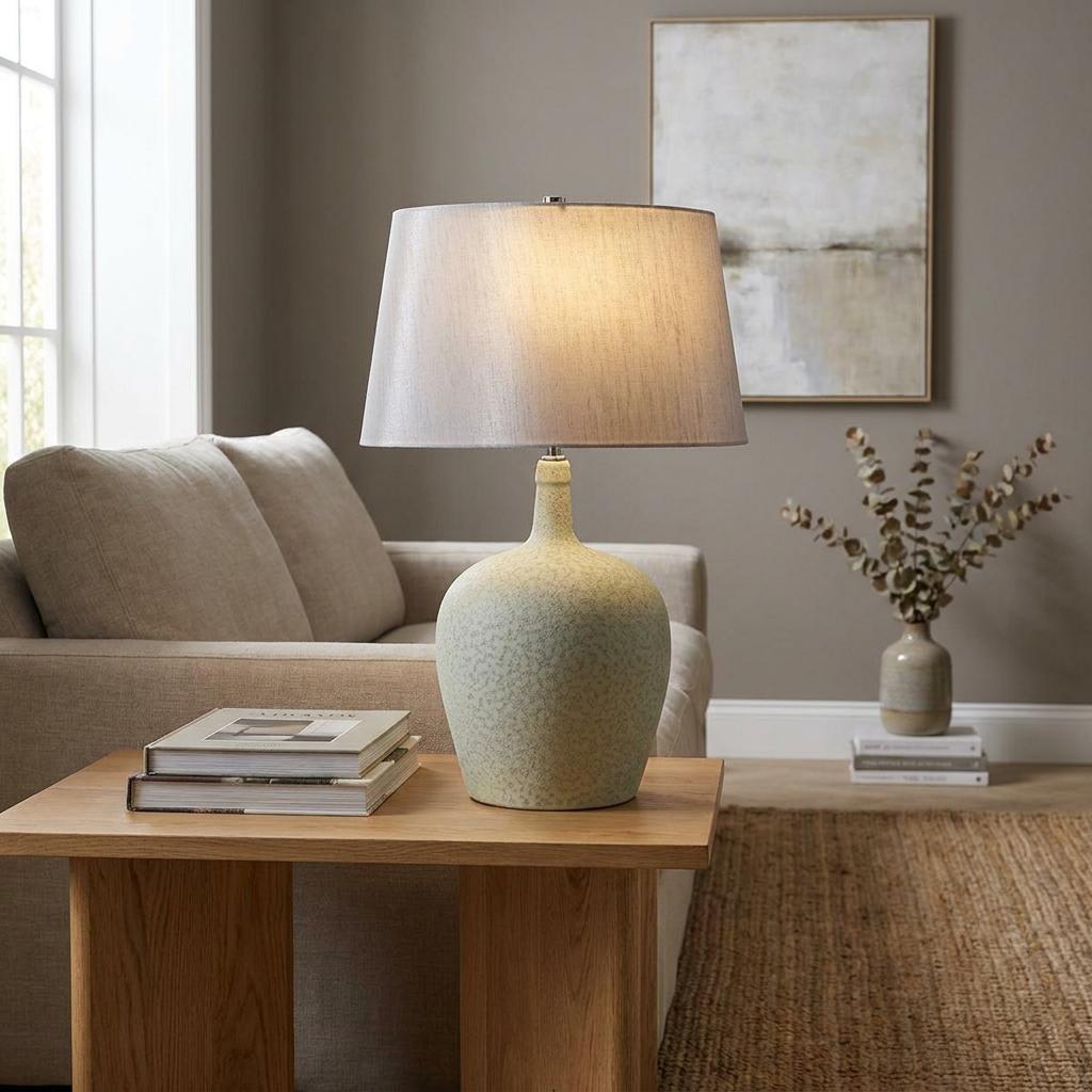 Sage green ceramic table lamp with a grey fabric shade on a wooden side table in a modern living room.