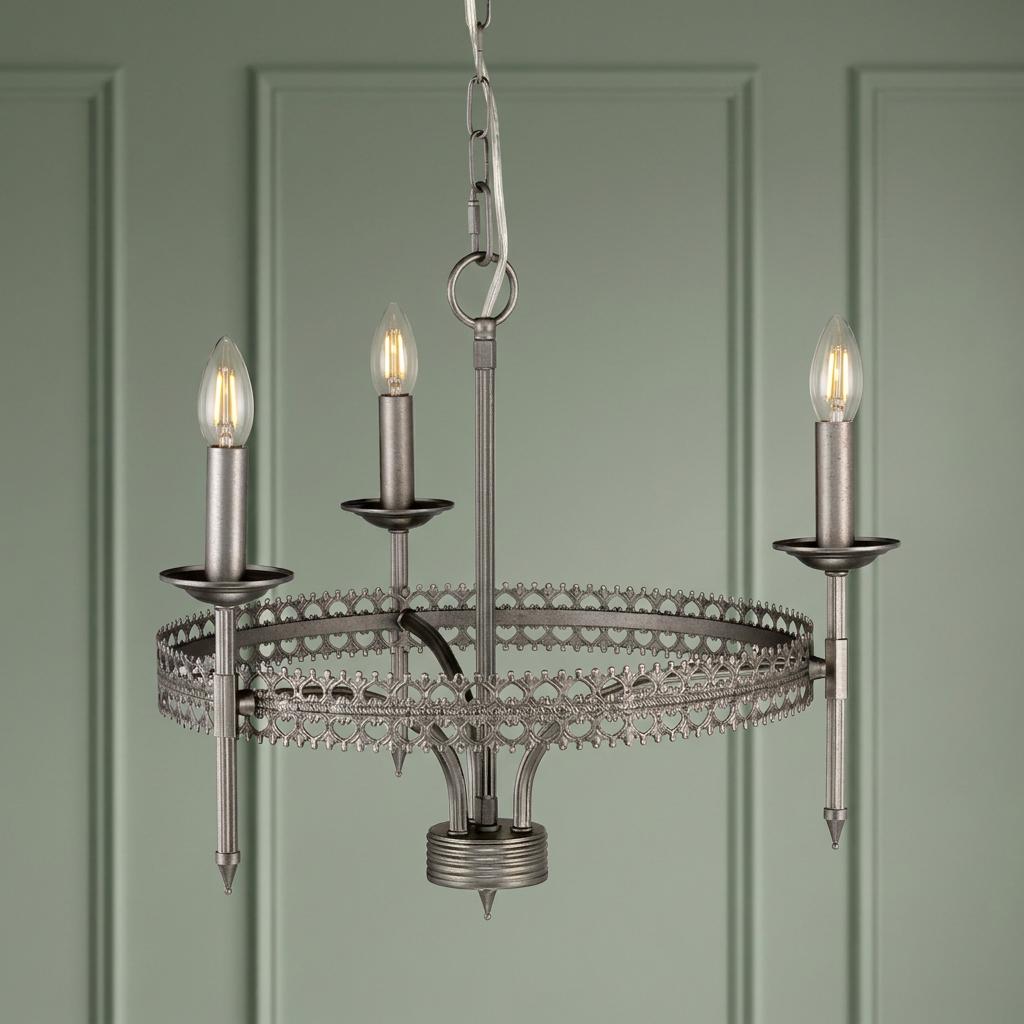 Silver 3-light crown chandelier with intricate metal scrollwork, hanging in front of a green paneled wall.