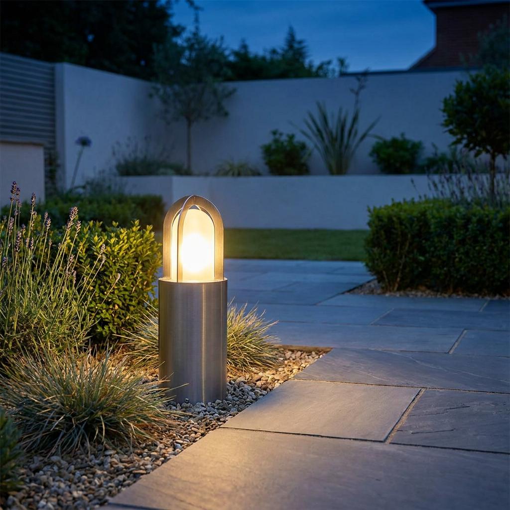 Silver Elstead Paignton bollard light with a warm glow, set in a gravel garden border next to a stone patio at night.