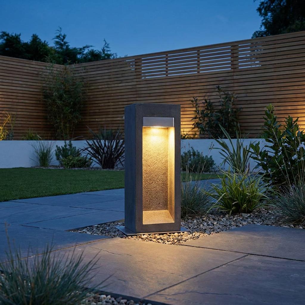 A dark grey concrete LED bollard light illuminates a landscaped garden path at night near a wooden fence.