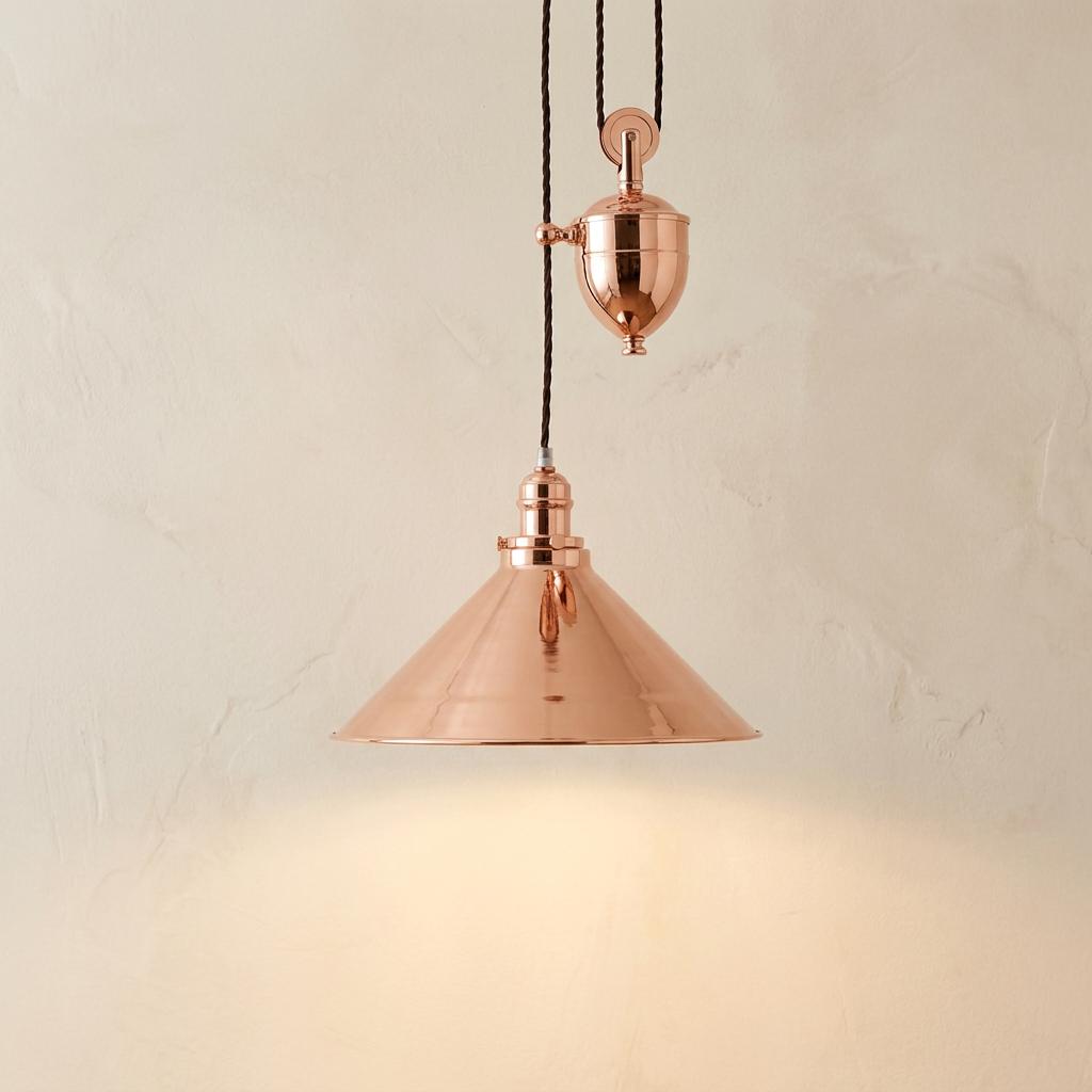 Polished copper rise and fall pendant light with a conical shade and pulley mechanism against a textured wall.