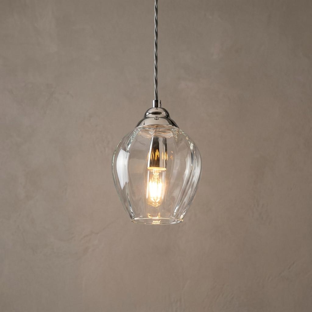 Polished nickel pendant light with a fluted clear glass shade and a glowing bulb against a neutral wall.