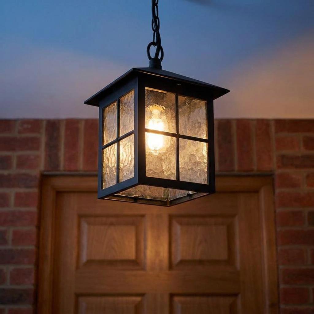 Black lantern-style outdoor pendant light with textured glass panels, lit in front of a wooden door and brick wall.