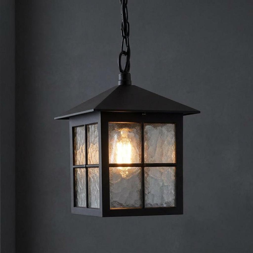 Black lantern-style outdoor pendant light with textured glass panes and a warm glowing bulb against a dark wall.