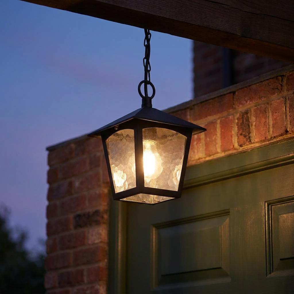 A black lantern-style pendant light with glowing textured glass hangs from a chain outside a brick building at dusk.