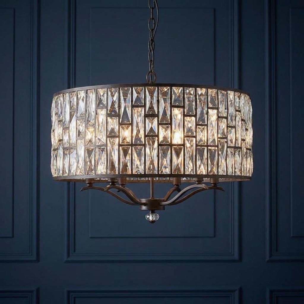 Bronze drum pendant light with clear crystal panels, lit and hanging against a dark blue paneled wall.