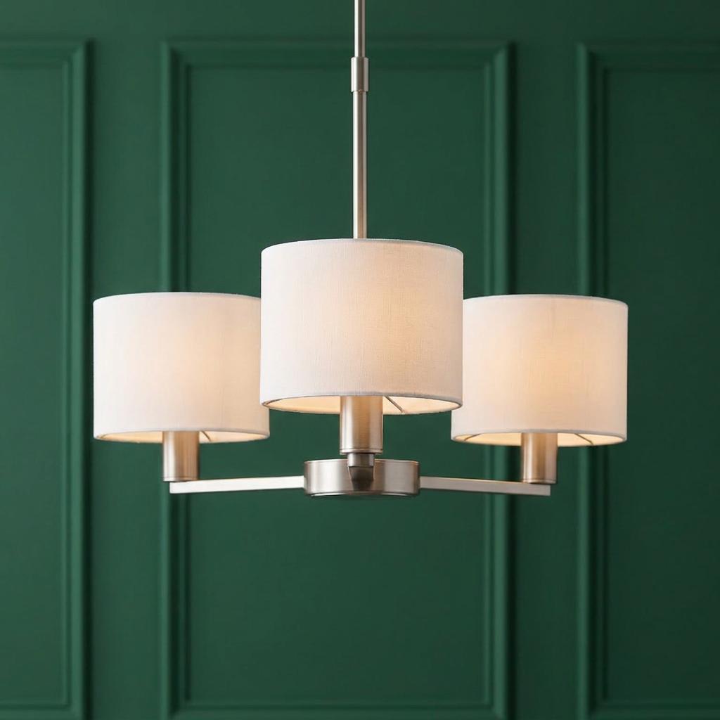 Three-light satin nickel ceiling pendant with white drum shades against a dark green paneled wall.