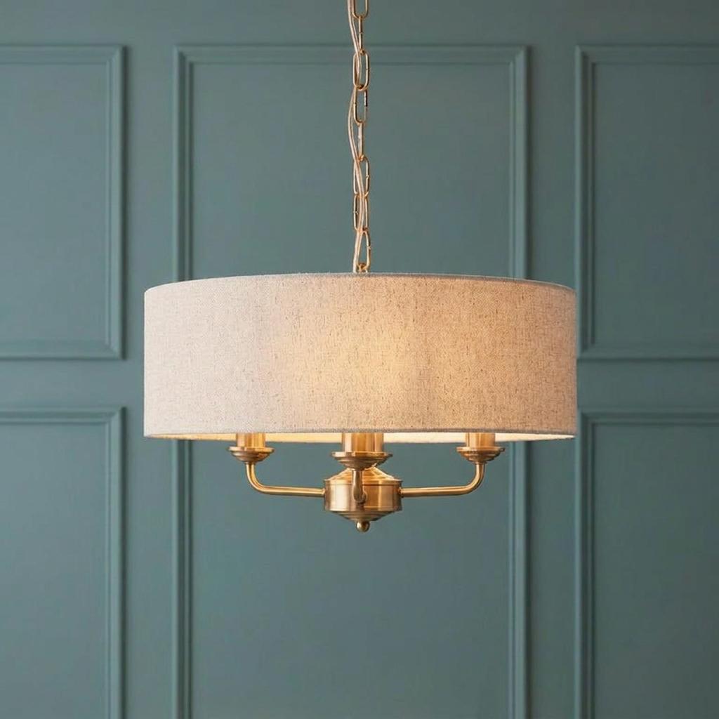 Aged brass three-light pendant with a natural linen drum shade against a teal paneled wall.