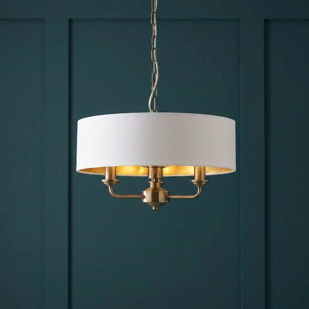 Antique brass 3-light pendant with a white drum shade hanging against a dark teal paneled wall.