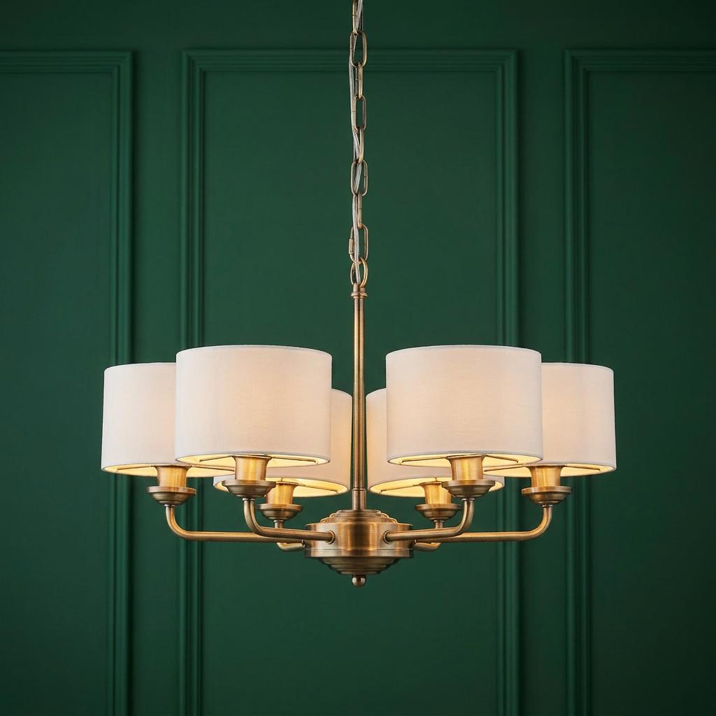 Antique brass 6-arm pendant chandelier with white fabric shades against a dark green paneled wall.