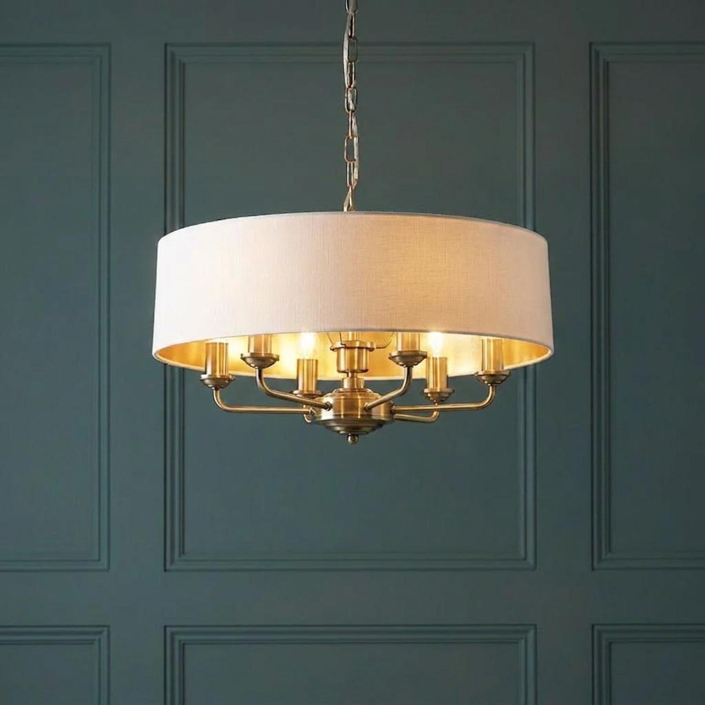 Antique brass 6-light pendant with a white drum shade and gold interior against a dark teal paneled wall.