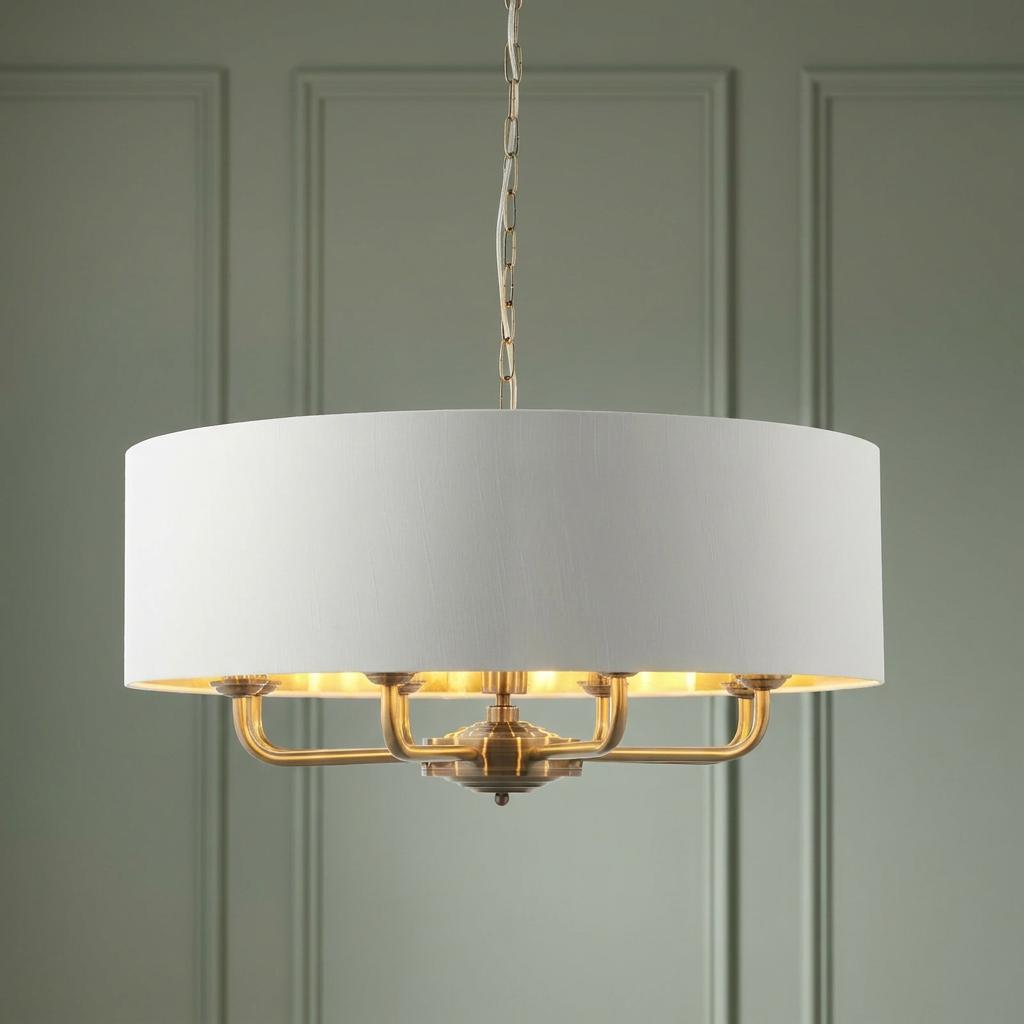 Antique brass 8-light pendant with a large white drum shade, illuminated against a sage green paneled wall.