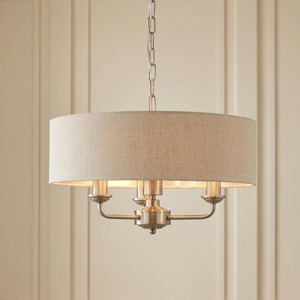 Brushed chrome 3-light pendant with a natural linen drum shade, hung by a chain against a cream wall.