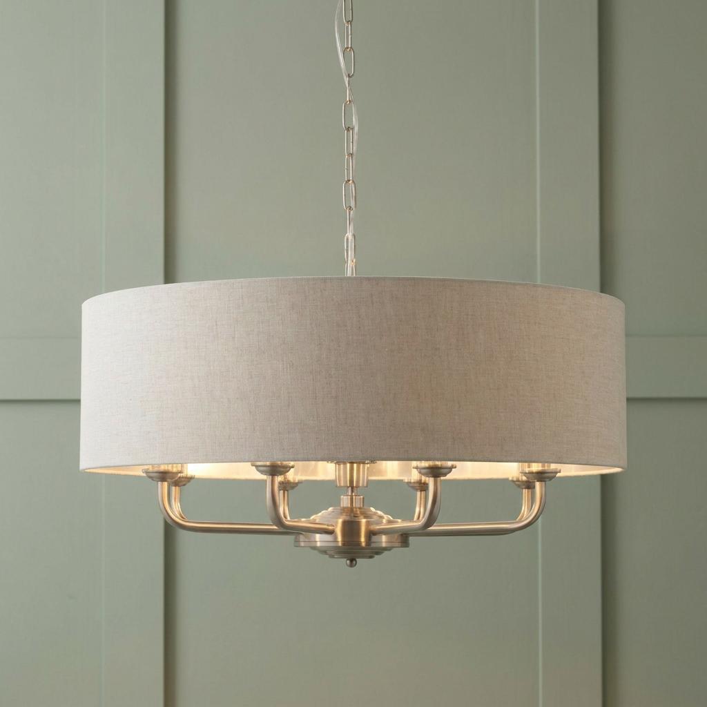 Brushed chrome 8-light pendant with a natural linen drum shade against a sage green paneled wall.