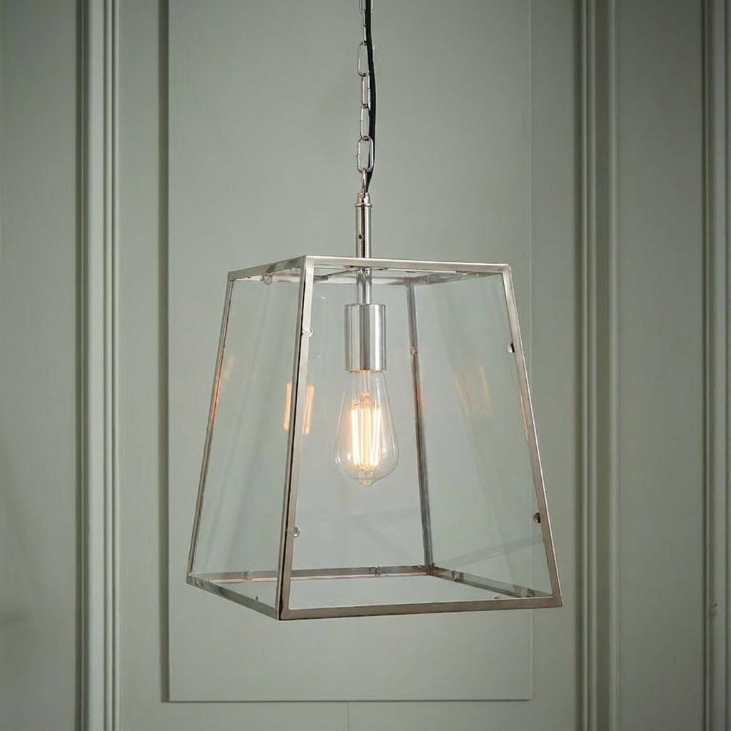 Polished nickel lantern pendant light with clear glass and an exposed Edison bulb, hanging against a paneled wall.
