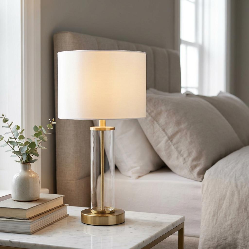 Modern gold and glass table lamp with a white drum shade on a marble bedside table in a neutral bedroom.