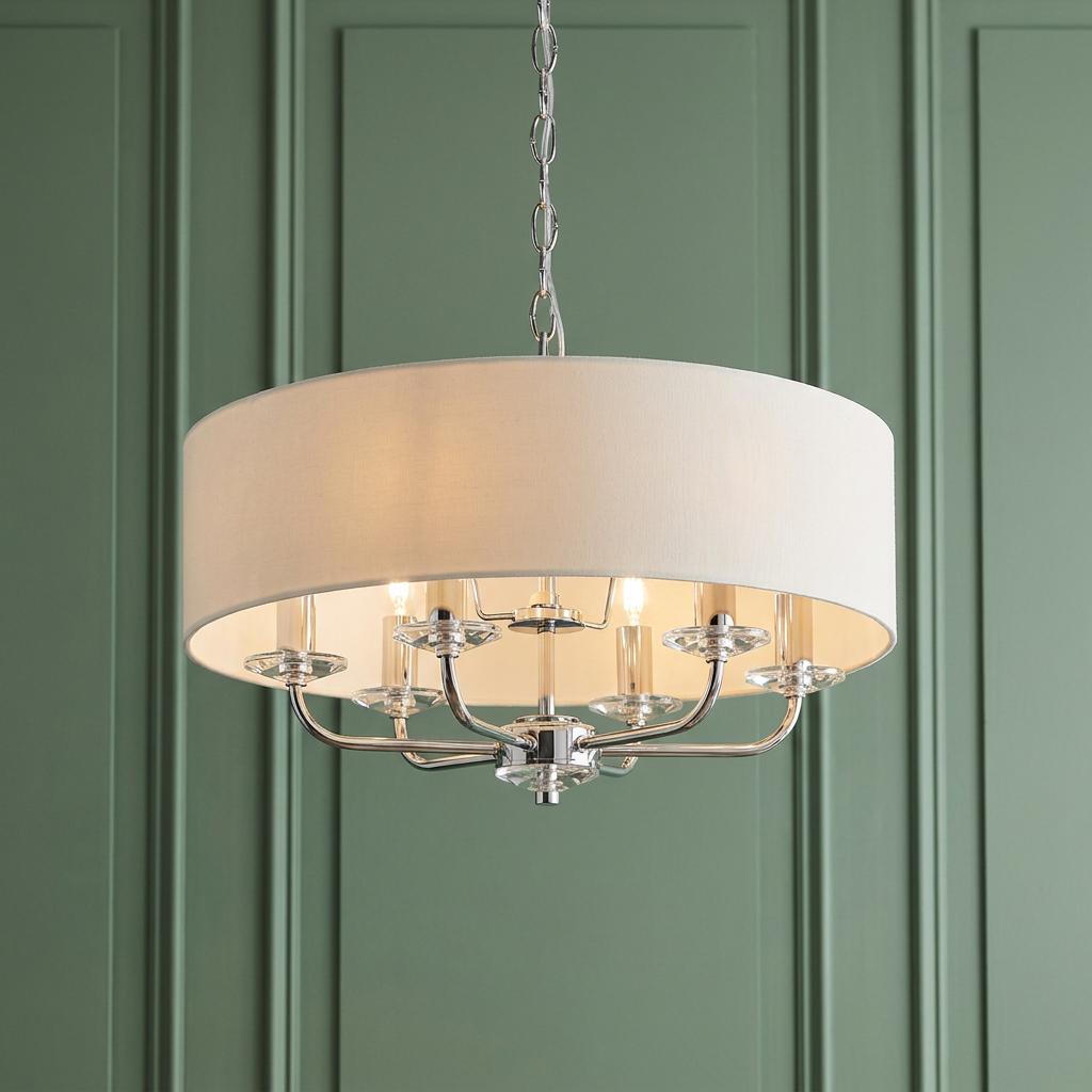 A six-arm bright nickel pendant light with a cream drum shade and crystal details against a green paneled wall.