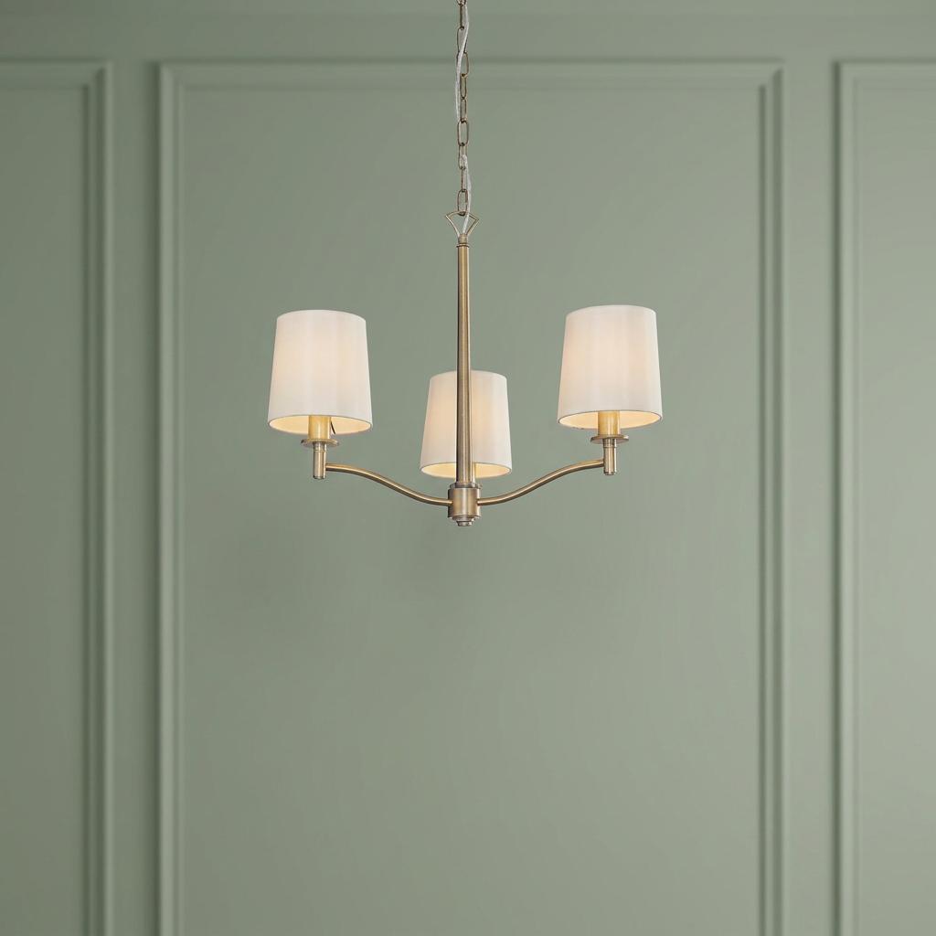 Endon Ortona 3-light antique brass ceiling pendant with white fabric shades against a sage green paneled wall.