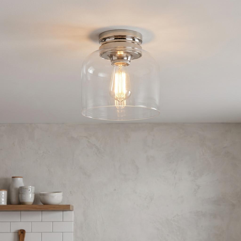 Polished nickel flush mount ceiling light with a clear glass bell shade and glowing Edison bulb on a white ceiling.