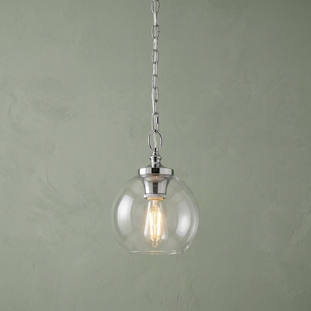 A Feiss Tabby polished nickel pendant light with a clear glass globe and glowing bulb against a green wall.
