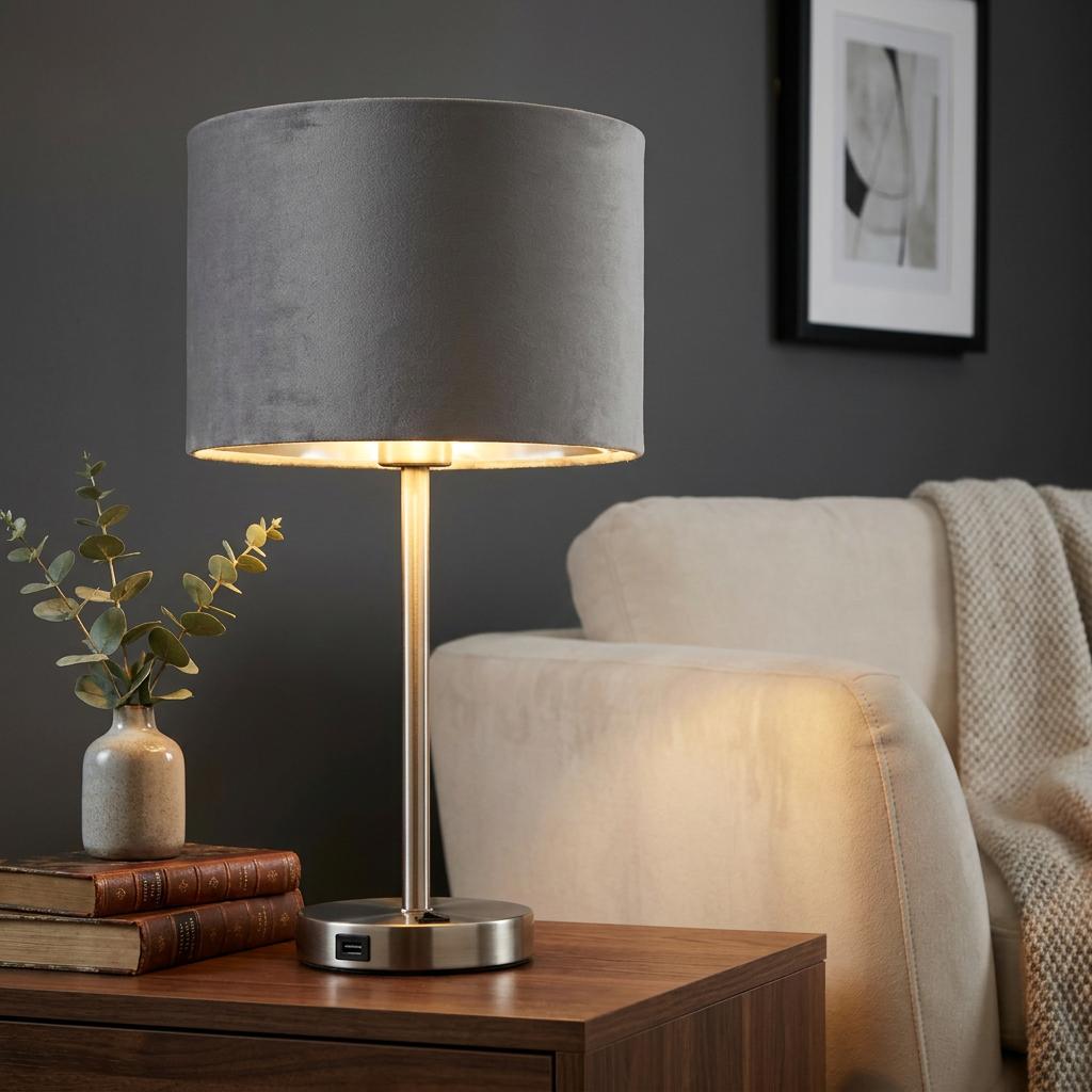 Illuminated satin nickel table lamp with a grey velvet shade on a wooden side table in a modern living room.