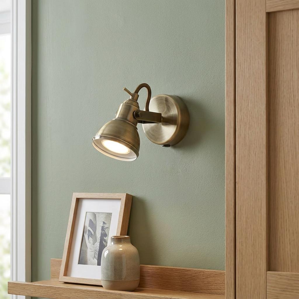 Antique brass adjustable wall spotlight mounted on a green wall above a wooden shelf with decor.