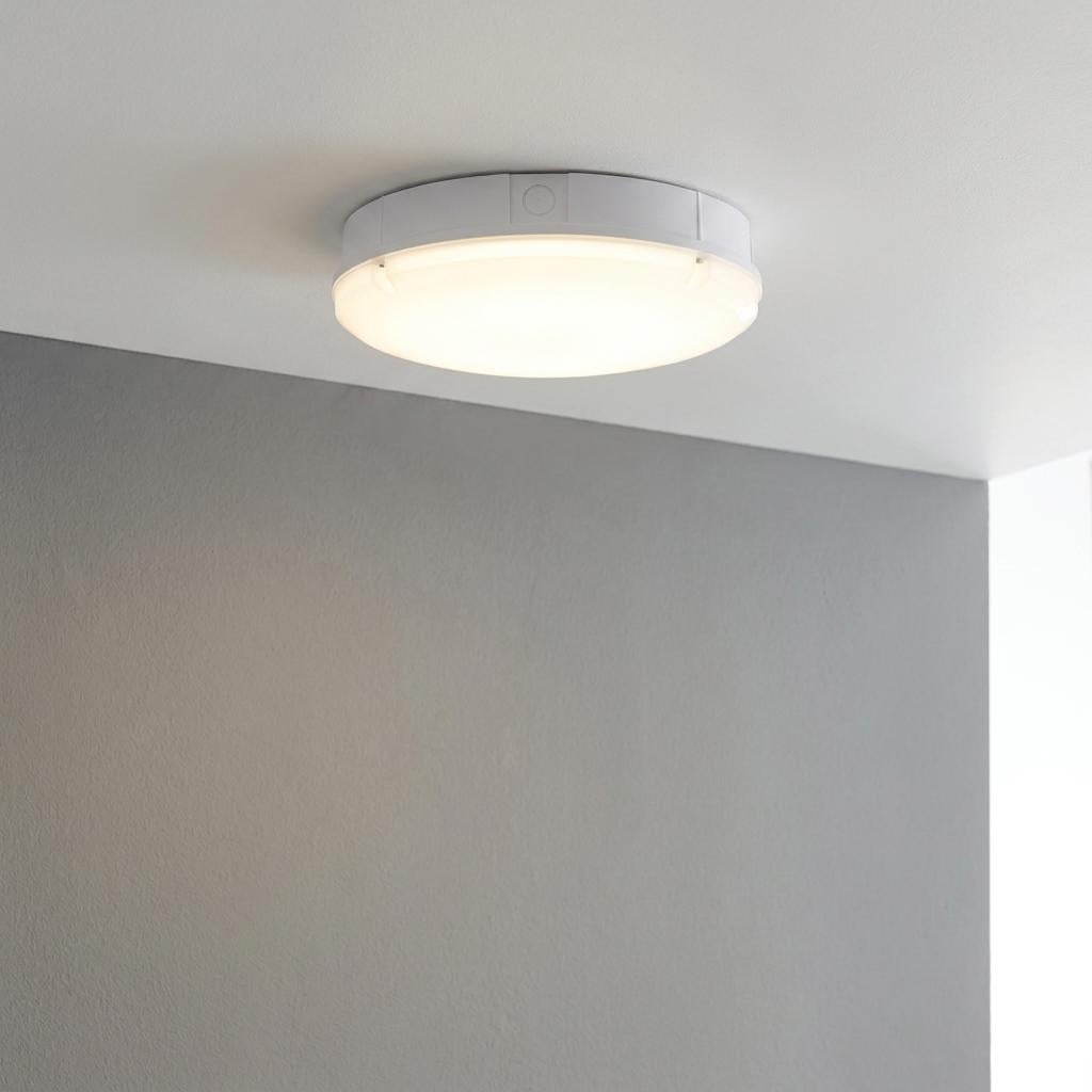 Round white bulkhead light glowing warmly while mounted on a white ceiling next to a grey wall.