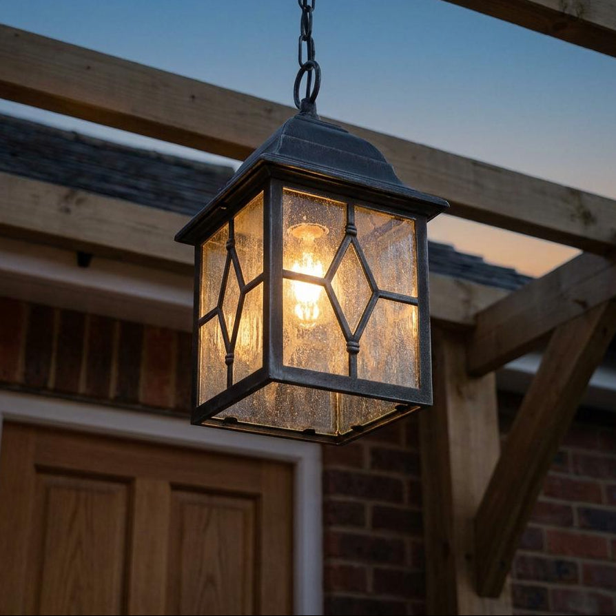 Black silver outdoor pendant light with leaded glass panels hanging from a wooden pergola at dusk.