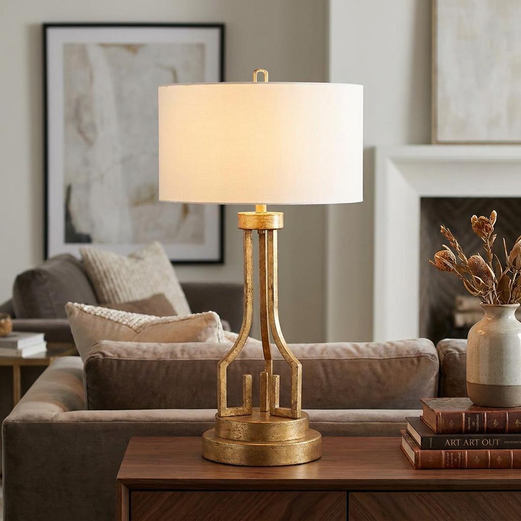Distressed gold table lamp with a geometric base and white drum shade, positioned on a wooden table in a living room.