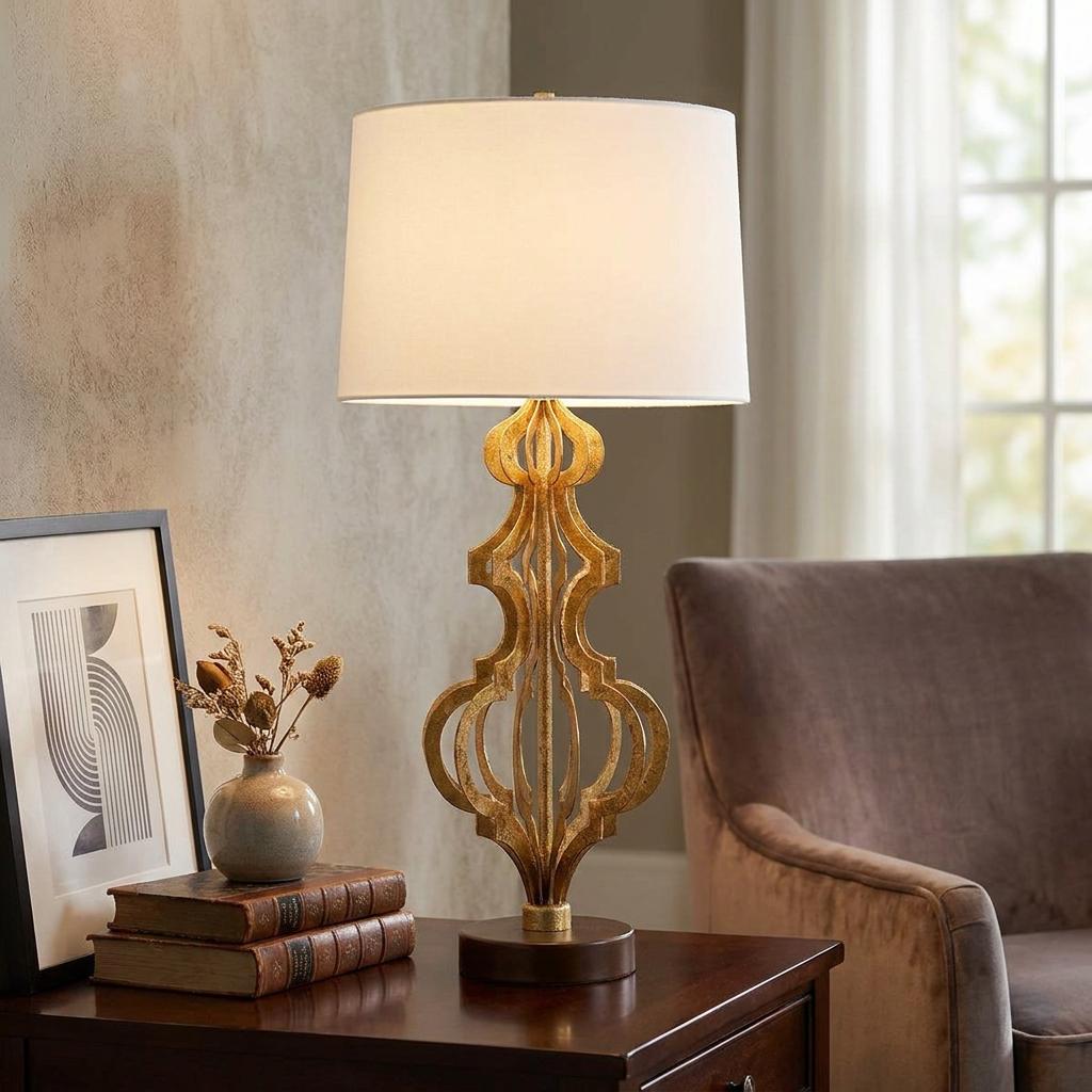 Gilded gold table lamp with a sculptural geometric base and white drum shade on a wooden side table.