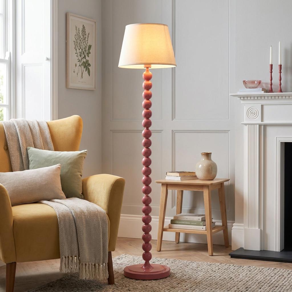 A pink bobble-stem floor lamp with a white shade standing by a yellow armchair in a neutrally decorated living room.