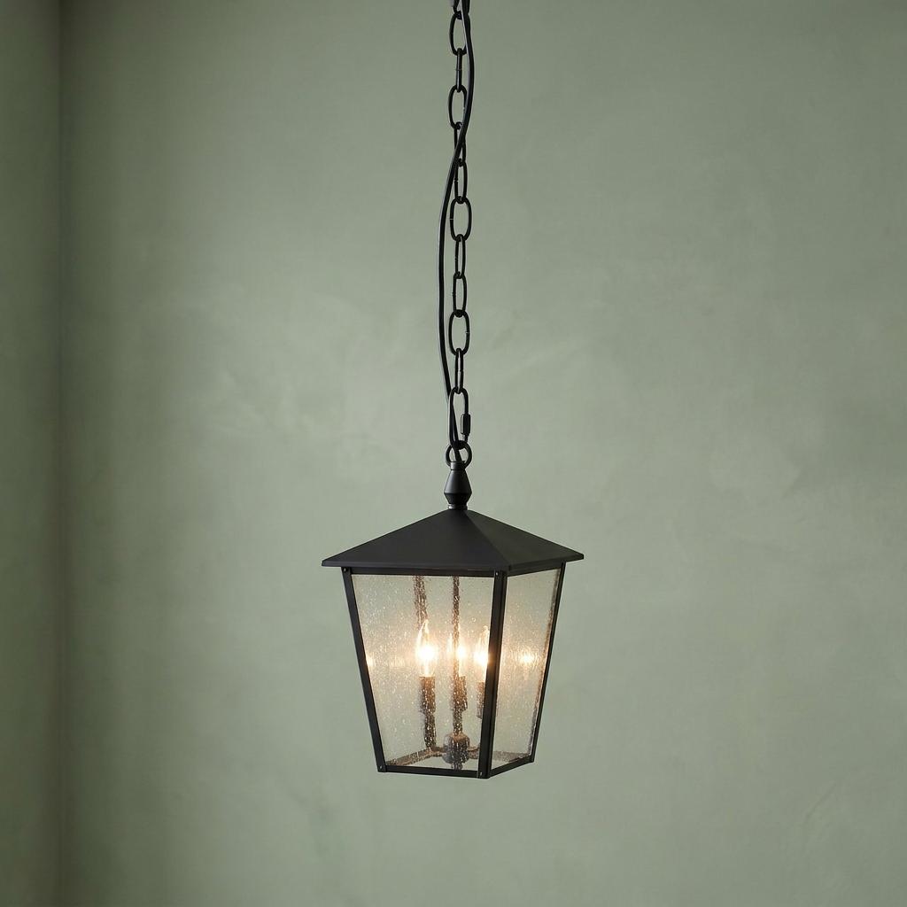 Black outdoor chain lantern with seeded glass and three illuminated candle lights against a pale green wall.