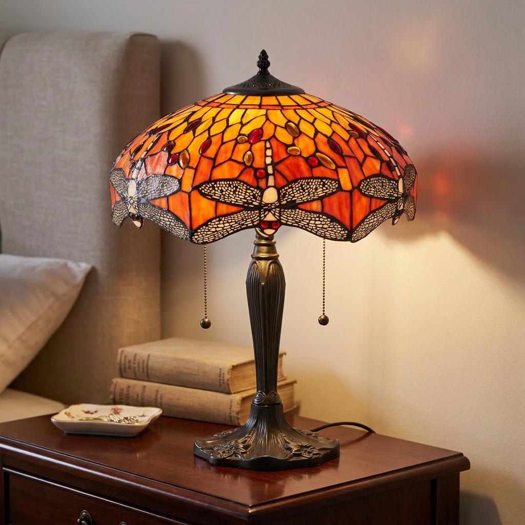 A glowing Tiffany table lamp with an orange dragonfly shade on a dark wood nightstand next to a bed.