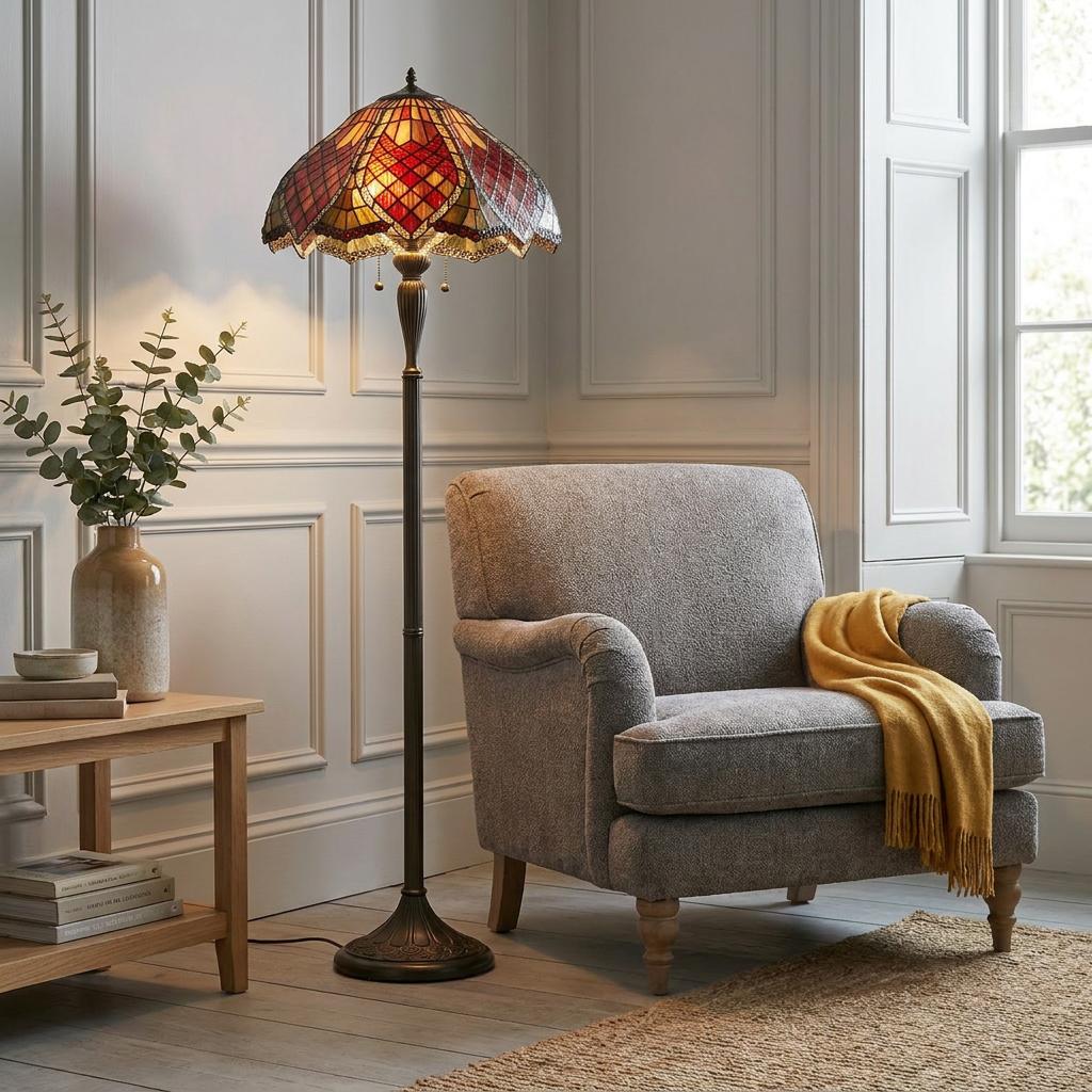 Interiors 1900 Orsino Tiffany floor lamp with a red and amber stained glass shade standing next to a grey armchair.
