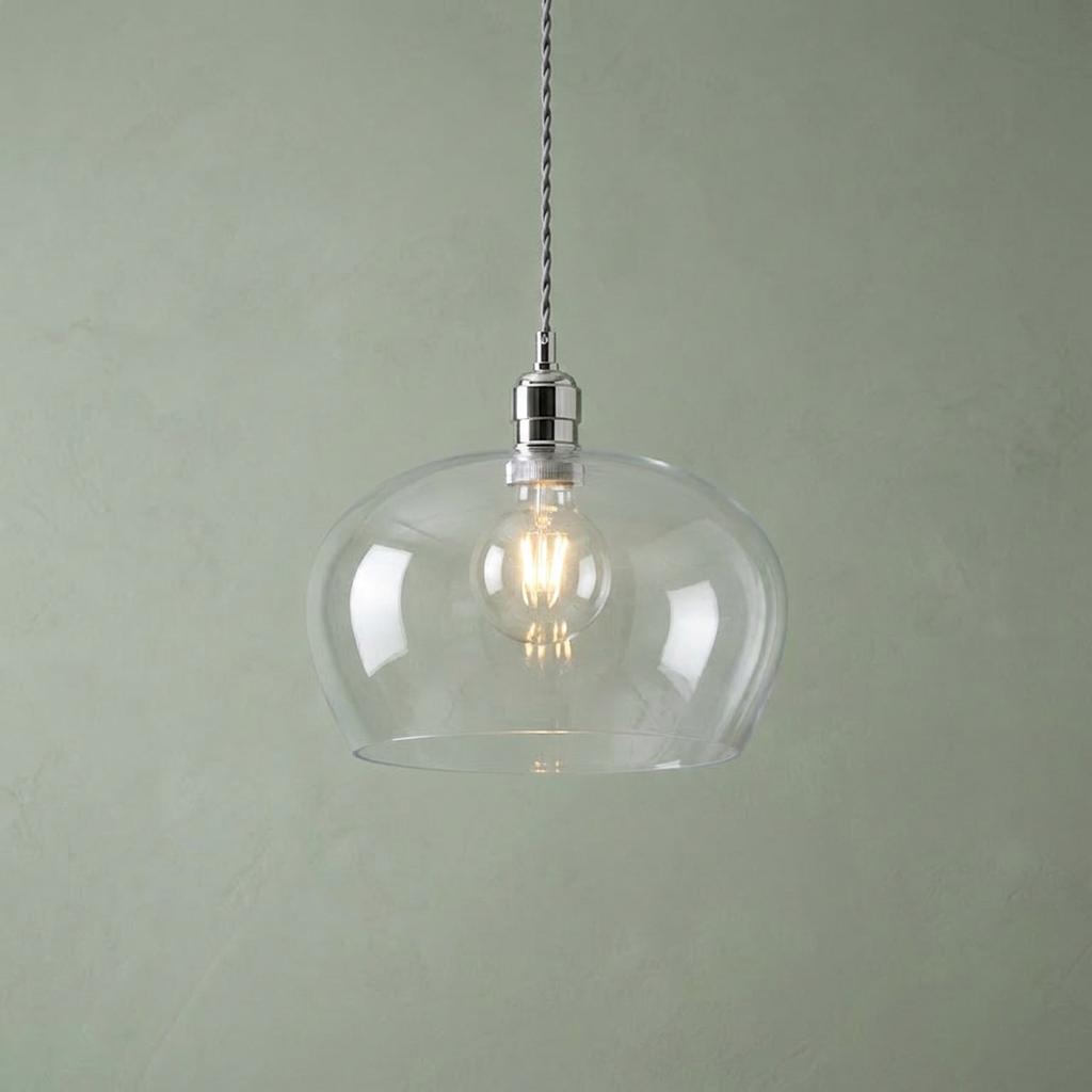 Nickel pendant light with a clear glass bowl shade and glowing bulb against a sage green textured wall.