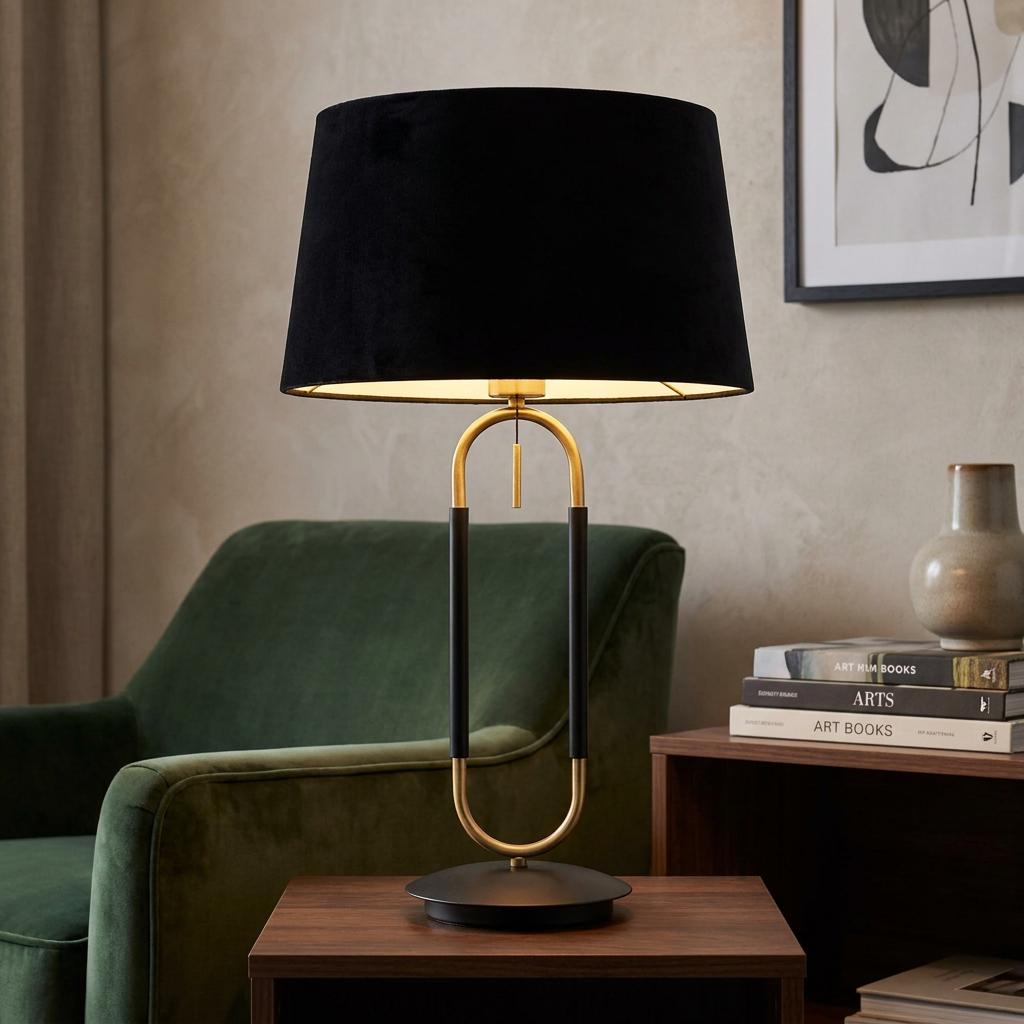 Brass and black table lamp with a black velvet shade on a wooden side table next to a green velvet armchair.