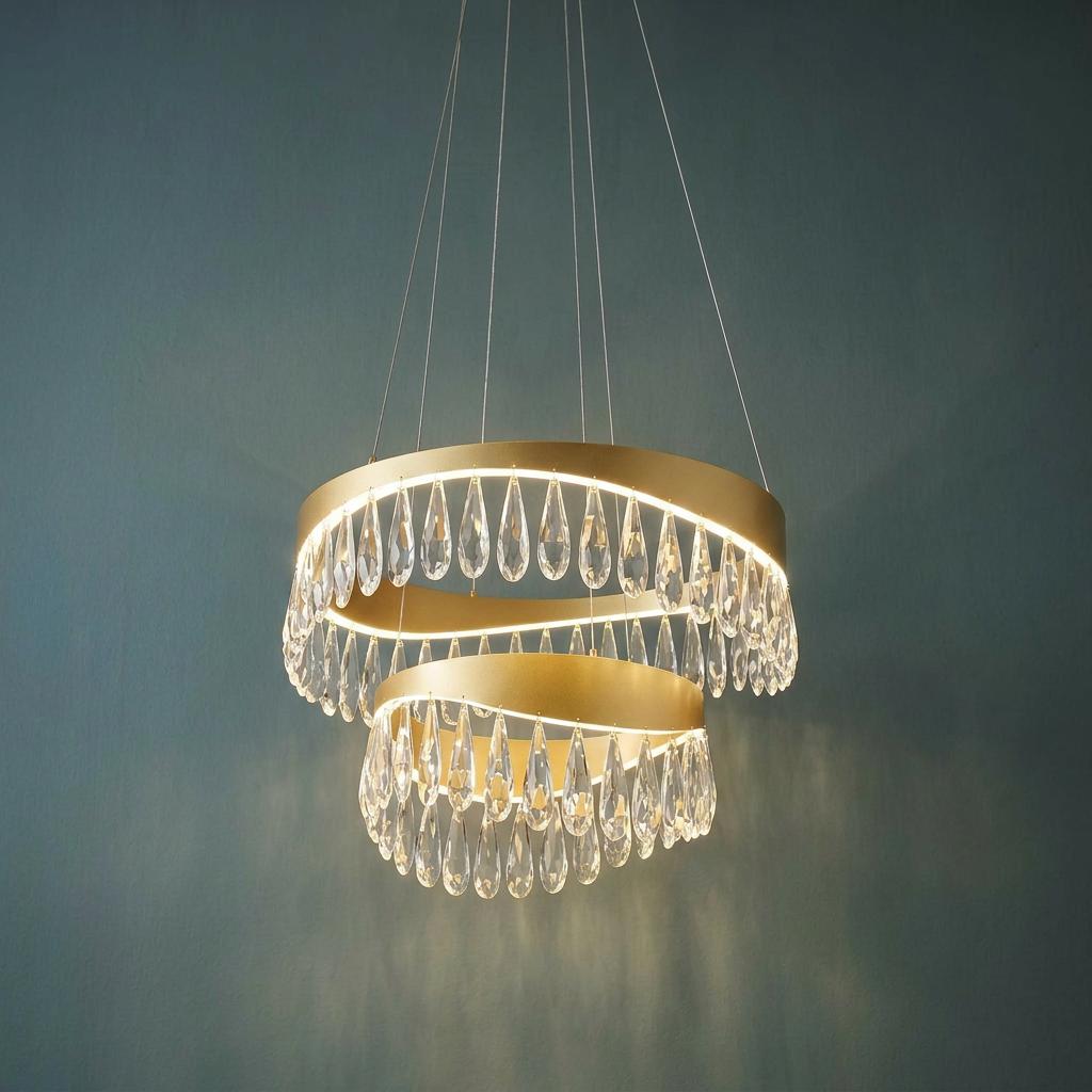 Two-tier gold pendant light with glowing LED bands and teardrop crystal droplets against a blue wall.
