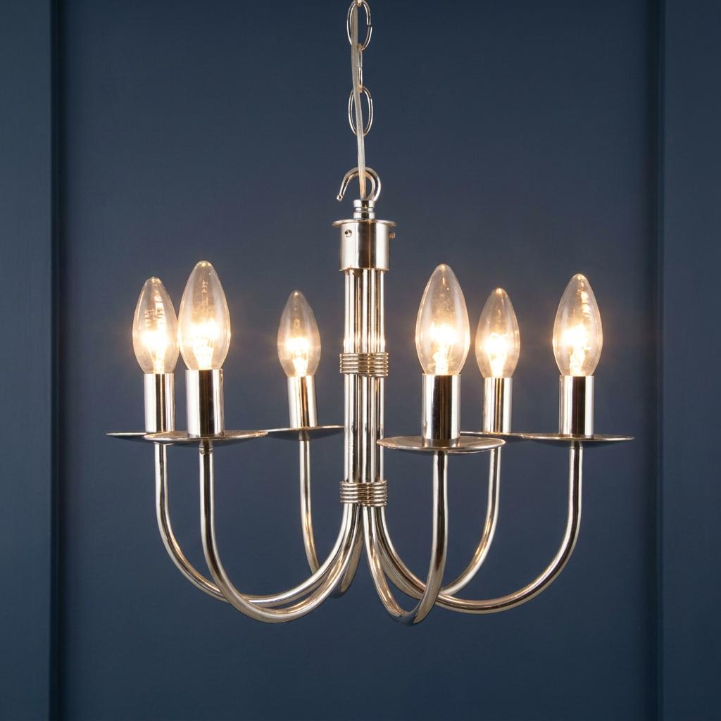 A polished stainless steel six-light chandelier with illuminated candle bulbs against a dark blue panelled wall.