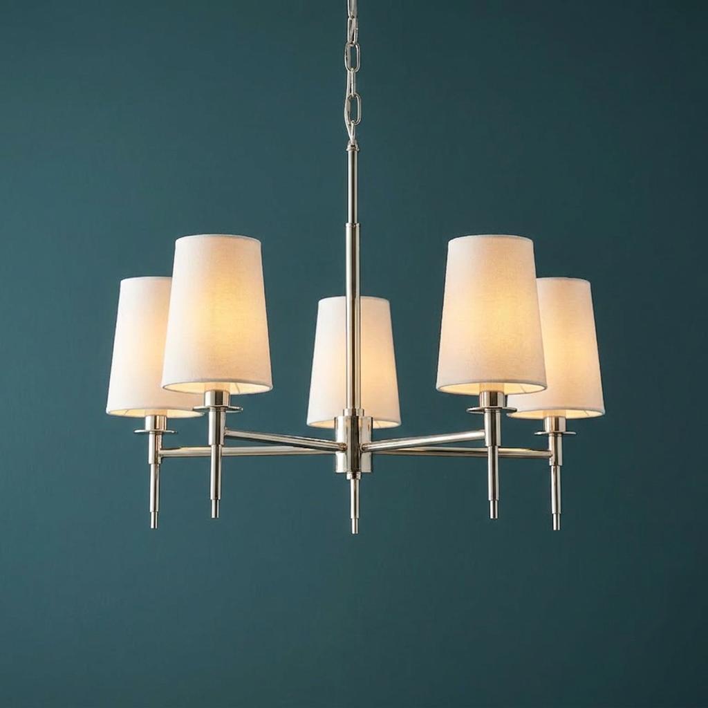 Five-light polished nickel pendant with white tapered fabric shades glowing against a deep teal wall.
