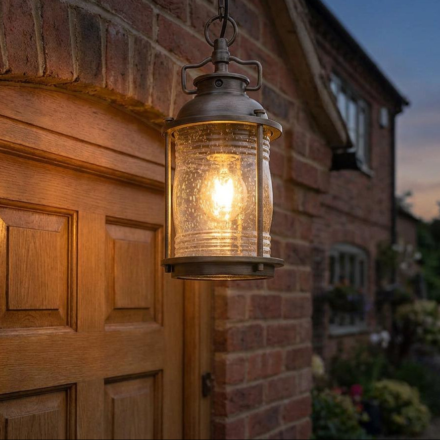 Bronze hanging lantern with seeded glass glowing warmly against a brick exterior and wooden door at twilight.