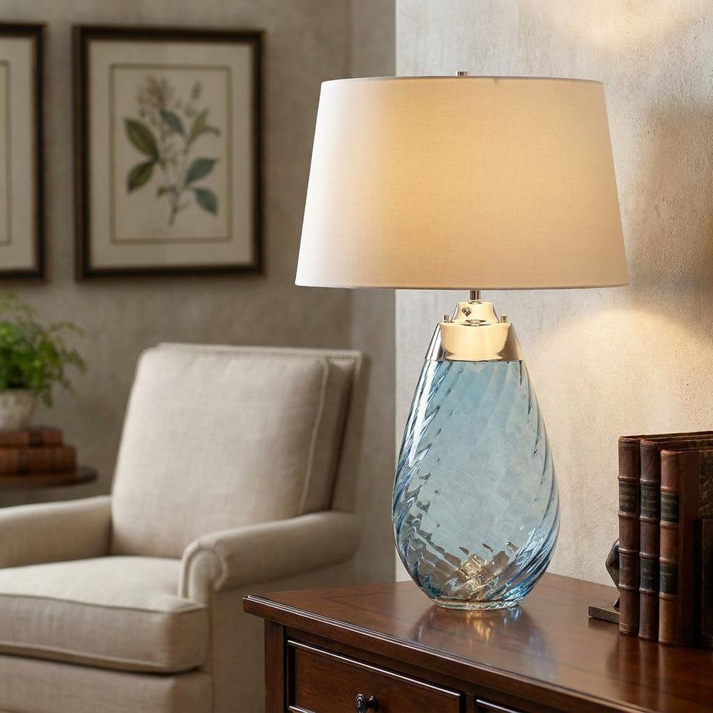 Blue glass table lamp with off-white shade on a wooden table in a living room setting.