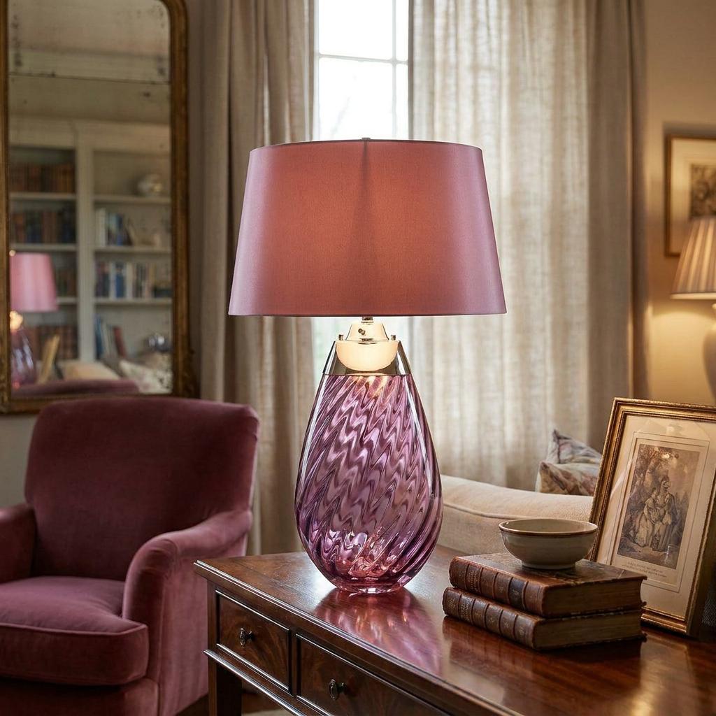 Plum glass table lamp with a matching shade sits on a wooden desk in a cozy living room setting.