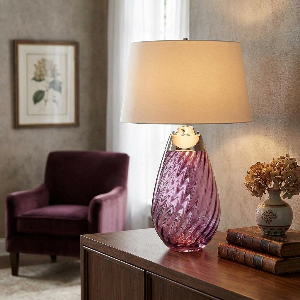 Plum glass table lamp with a twisted base and cream shade lit on a wooden sideboard in a living room.