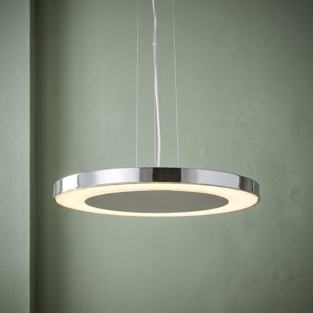 A modern chrome LED disc pendant light with a glowing inner ring, suspended against a sage green wall.