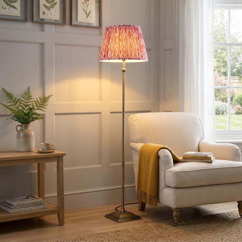 Lille brass floor lamp with coral pink ikat shade beside a beige armchair in a warmly lit, paneled living room.
