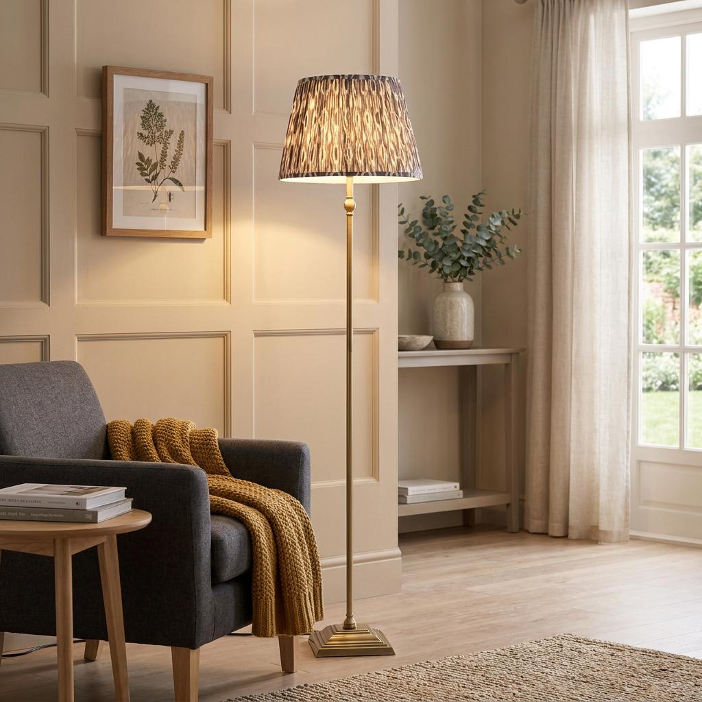 Lille brass floor lamp with a grey ikat shade standing next to a grey armchair in a neutral-toned living room.