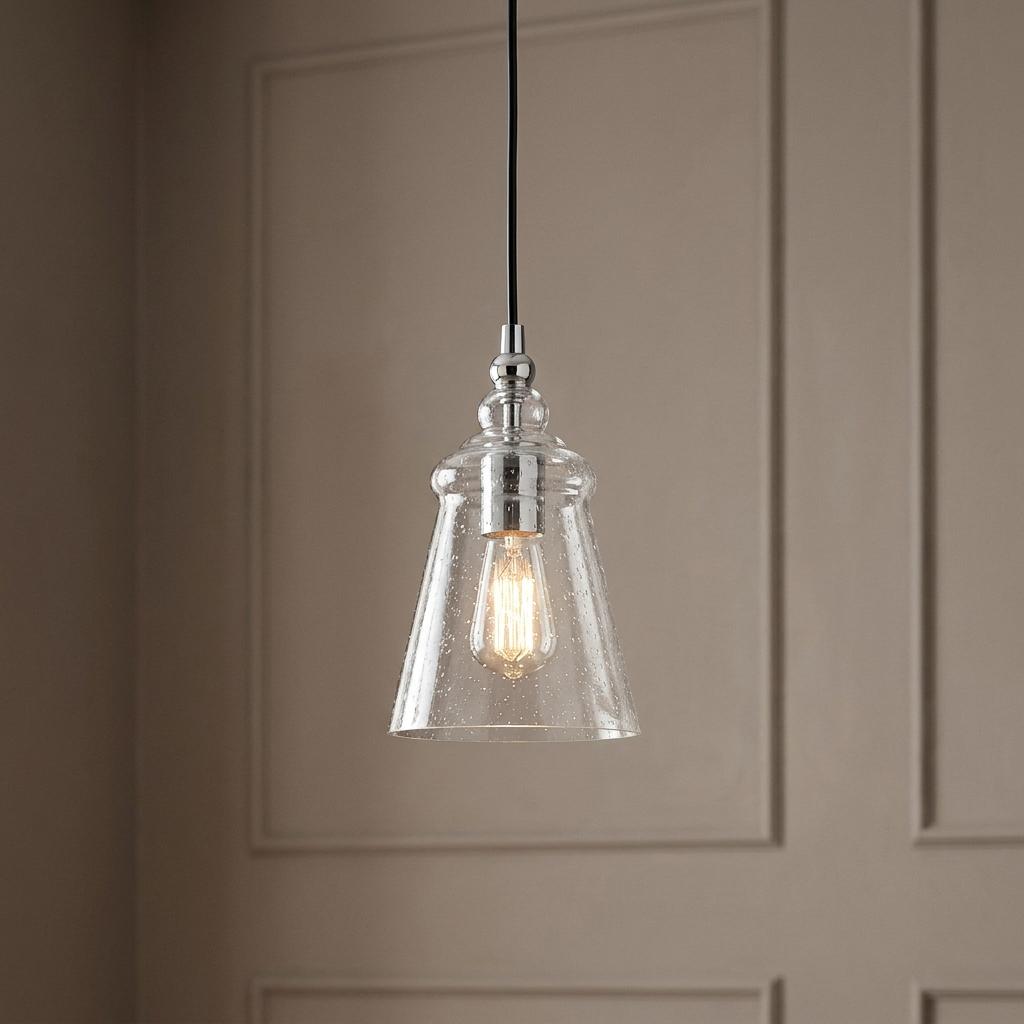 Mini chrome pendant light with a tapered seeded glass shade and glowing filament bulb against a paneled wall.