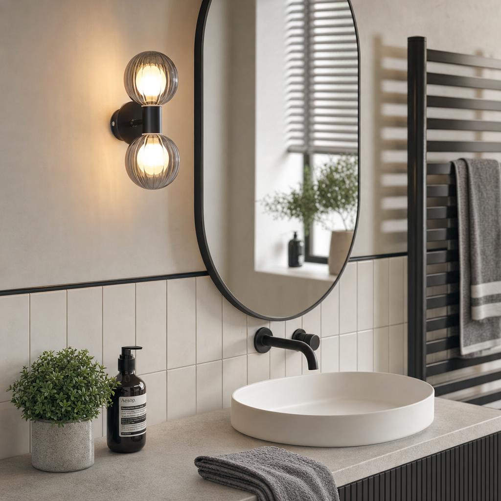 Matt black bathroom wall light with two ribbed glass globes mounted next to an oval mirror above a white vessel sink.