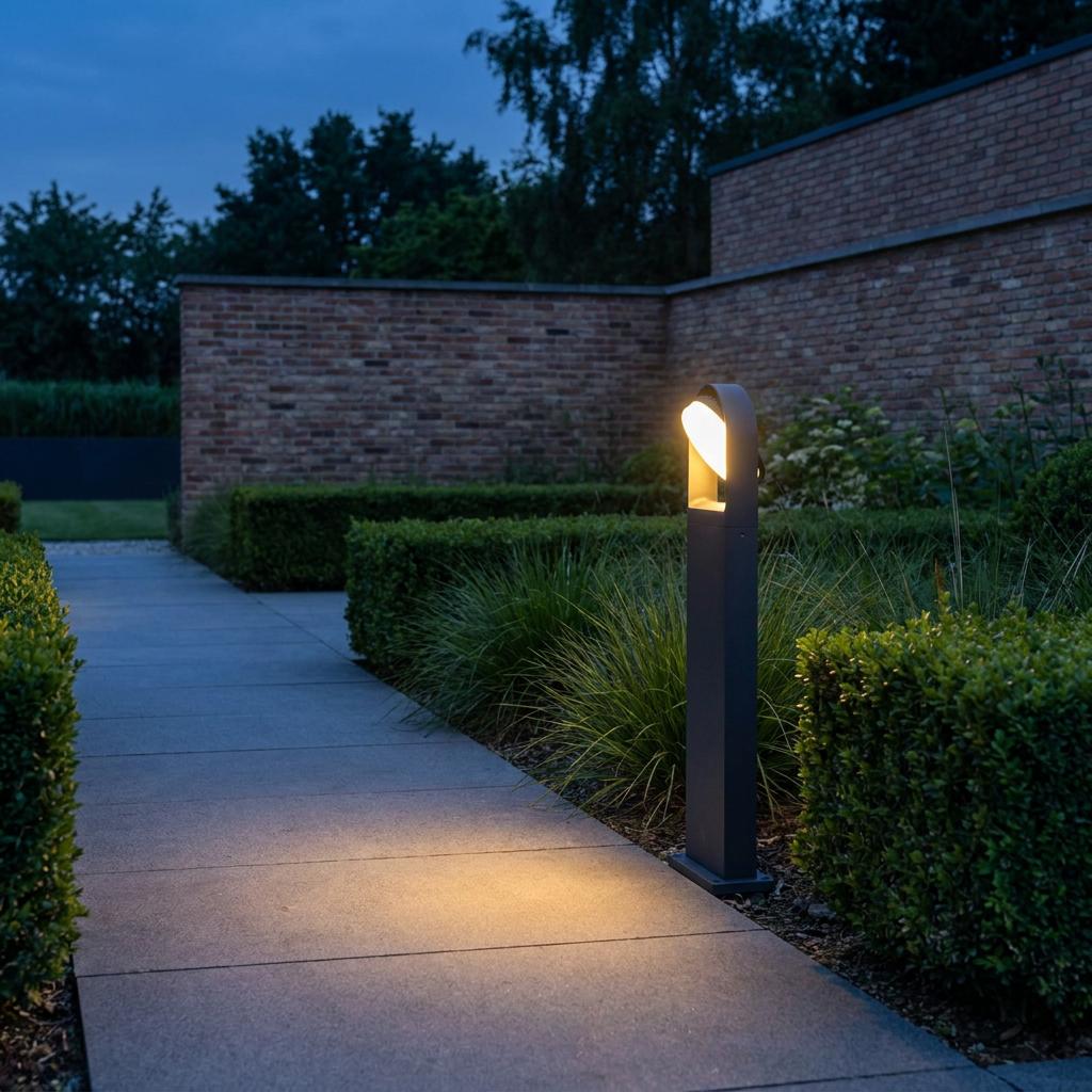 A grey Lutec Fele bollard light illuminating a stone garden path at dusk next to manicured hedges.